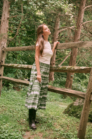 A person wearing the Roller Rabbit Hayam Carlisa Skirt in an olive and white print, paired with a cream knit top and brown boots, stands against a plain white background.