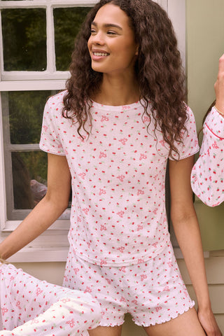 A smiling woman with curly hair stands outdoors near a window, wearing the Roller Rabbit Mini Monkey Blossom Pointelle Classic Tee—a white short-sleeve top featuring a delicate pink floral print.
