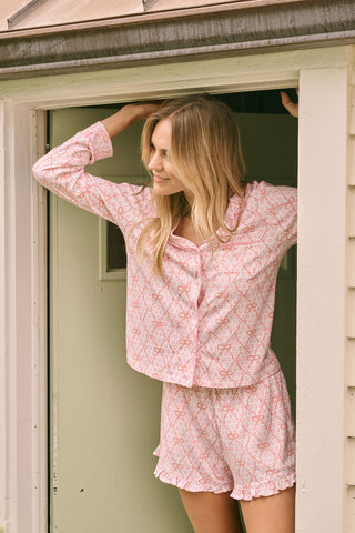 Wearing Roller Rabbit’s Lasso Bows Hayden Pajamas in pastel pink, a woman pairs the two-piece set with brown cowboy boots as she stands by a light-colored house door, smiling and resting one arm above her head while bending one leg with her foot on a step.