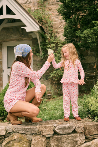 A young girl stands smiling, wearing the Roller Rabbit Kids Apple Bloom Elle Set—a pink girls’ loungewear outfit featuring an apple bloom print and a ruffled hem. She has long dark hair and is wearing light pink shoes, set against a plain white background.