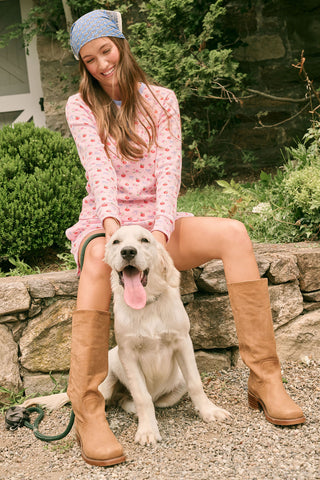 A young girl stands smiling, wearing the Roller Rabbit Kids Apple Bloom Elle Set—a pink girls’ loungewear outfit featuring an apple bloom print and a ruffled hem. She has long dark hair and is wearing light pink shoes, set against a plain white background.