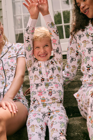 A smiling child in Roller Rabbit’s Kids Boo Crew Pajamas, crafted from soft Pima cotton, sits with arms raised on outdoor steps among women in coordinating patterned pajamas, all sharing a cheerful and relaxed moment together.