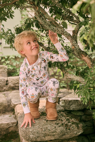 A smiling child in Roller Rabbit’s Kids Boo Crew Pajamas, crafted from soft Pima cotton, sits with arms raised on outdoor steps among women in coordinating patterned pajamas, all sharing a cheerful and relaxed moment together.