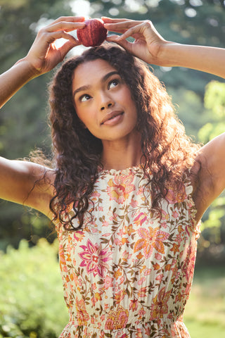 A woman in the Priya Floral Milani Dress by Roller Rabbit and boots stands on grass, holding a hat filled with red apples. Trees and sunlight fill the background.