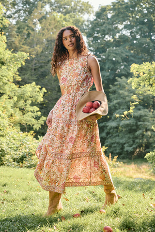 A woman in the Priya Floral Milani Dress by Roller Rabbit and boots stands on grass, holding a hat filled with red apples. Trees and sunlight fill the background.