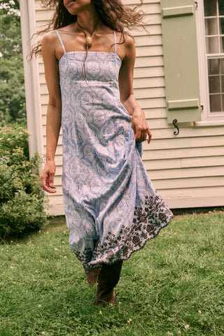 A woman wearing the Roller Rabbit Lakai Asher Dress—a light blue patterned cotton batiste dress with dark lace trim—walks on grass near a cream-colored house with green shutters, partially visible.