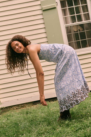 A woman wearing the Roller Rabbit Lakai Asher Dress—a light blue patterned cotton batiste dress with dark lace trim—walks on grass near a cream-colored house with green shutters, partially visible.