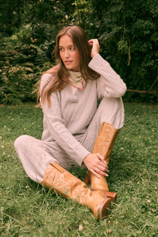 A woman wearing a light gray Cable Knots Rocio Sweater by Roller Rabbit, featuring a textured knit and V-neck, paired with matching knit pants, stands against a plain white background.