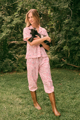 Wearing the Charlie Liza Set by Roller Rabbit, a woman stands on the grass in brown boots, holding a small black animal, with green foliage in the background.