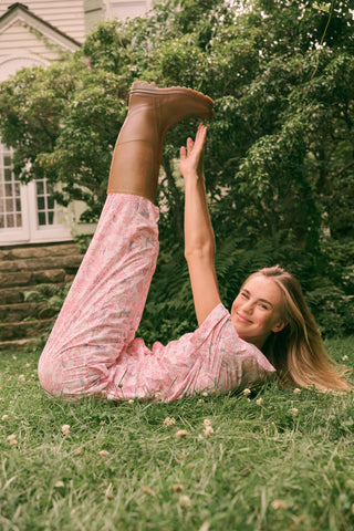 Wearing the Charlie Liza Set by Roller Rabbit, a woman stands on the grass in brown boots, holding a small black animal, with green foliage in the background.
