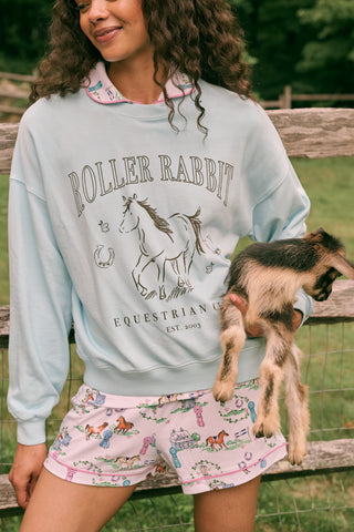 Wearing the Roller Rabbit Equestrian Club Phoebe Sweatshirt in baby blue, a woman pairs it with horse-patterned shorts as she holds a baby goat. She stands by a wooden fence with greenery in the background, smiling softly.
