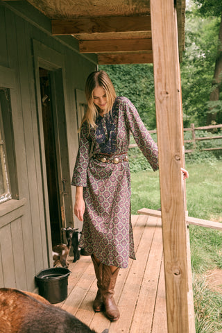 Wearing the Amelia Inez Long Kurta by Roller Rabbit, a woman with brown boots sits on the railing of a wooden porch, smiling, with a rustic building in the background.