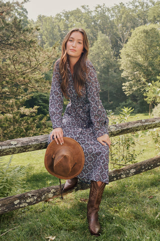 A smiling woman in the full-length Amelia Belted Darla Dress by Roller Rabbit sits on a wooden fence outdoors, holding a brown hat with trees and greenery in the background.