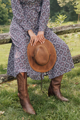 A smiling woman in the full-length Amelia Belted Darla Dress by Roller Rabbit sits on a wooden fence outdoors, holding a brown hat with trees and greenery in the background.