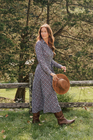A smiling woman in the full-length Amelia Belted Darla Dress by Roller Rabbit sits on a wooden fence outdoors, holding a brown hat with trees and greenery in the background.