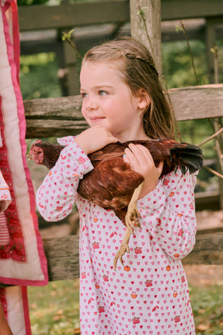 A young girl in a Roller Rabbit Kids Autumn Hearts Isa Dress and cowboy boots holds a brown chicken, standing outside next to an adult in a floral dress and boots, with grass and a wooden fence in the background.