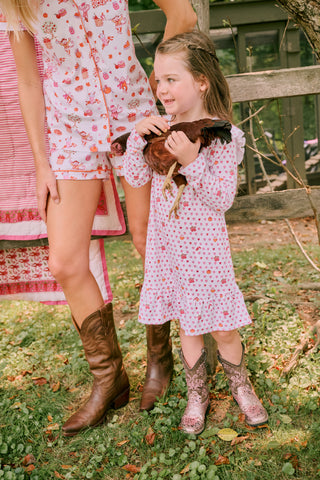 A young girl in a Roller Rabbit Kids Autumn Hearts Isa Dress and cowboy boots holds a brown chicken, standing outside next to an adult in a floral dress and boots, with grass and a wooden fence in the background.