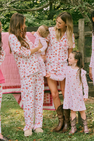 A woman smiles while wearing the Roller Rabbit Sweater Weather Polo Pajamas, a matching set of printed Pima PJs featuring colorful pink and orange cupcakes, flowers, and playful designs on a white background.