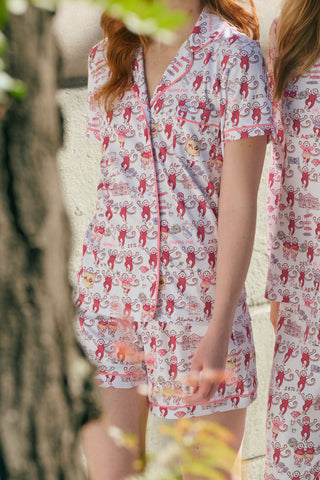 The Roller Rabbit Alpha Phi Monkey Polo Pajamas are a two-piece set crafted from soft Pima Cotton, featuring a white base adorned with playful red cartoon monkey prints and whimsical text. The short-sleeve top includes a front button placket, while the matching shorts feature an elastic waistband, all displayed on a plain background.