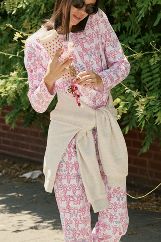 A woman wearing Roller Rabbit Gamma Phi Beta Monkey Pajamas—featuring a white and pink pattern with an elastic waistband—stands outdoors in front of greenery, holding a phone to her ear, a tumbler, and keys. A sweatshirt is tied around her waist, and she wears sunglasses.