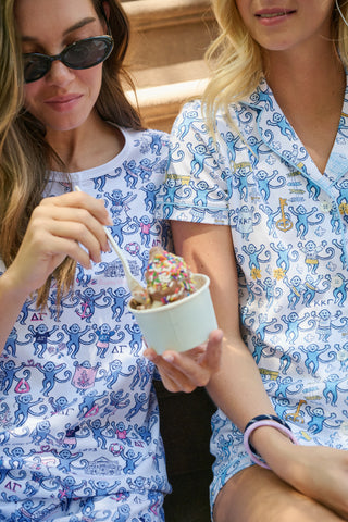 The Roller Rabbit Kappa Kappa Gamma Monkey Polo Pajamas feature a short-sleeve button-up pajama shirt with a front button placket and matching shorts with an elastic waistband, adorned with a playful blue monkey and yellow motif on a white background. Crafted from soft Pima cotton for extra comfort.