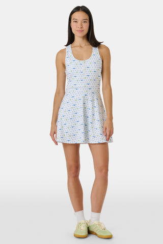 The Roller Rabbit Tennis Hearts Becky Active Dress is a sleeveless white active dress with a V-neckline and a flared skirt, adorned with an all-over pattern of small blue and green heart shapes. The dress is shown against a plain light gray background.