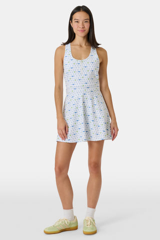 The Roller Rabbit Tennis Hearts Becky Active Dress is a sleeveless white active dress with a V-neckline and a flared skirt, adorned with an all-over pattern of small blue and green heart shapes. The dress is shown against a plain light gray background.