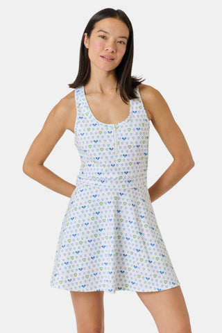 The Roller Rabbit Tennis Hearts Becky Active Dress is a sleeveless white active dress with a V-neckline and a flared skirt, adorned with an all-over pattern of small blue and green heart shapes. The dress is shown against a plain light gray background.