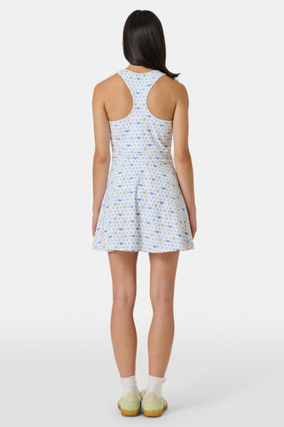 The Roller Rabbit Tennis Hearts Becky Active Dress is a sleeveless white active dress with a V-neckline and a flared skirt, adorned with an all-over pattern of small blue and green heart shapes. The dress is shown against a plain light gray background.