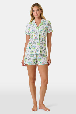 The Roller Rabbit Game, Set, Monkey Polo Pajamas is a two-piece set featuring a short-sleeve button-up shirt and matching shorts. Crafted from soft Pima Cotton, the white fabric is adorned with a playful cartoon animal and house print in green, blue, and yellow tones.