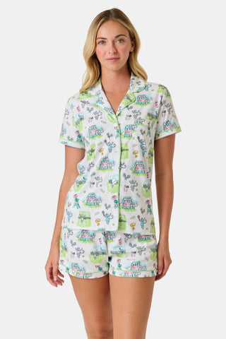 The Roller Rabbit Game, Set, Monkey Polo Pajamas is a two-piece set featuring a short-sleeve button-up shirt and matching shorts. Crafted from soft Pima Cotton, the white fabric is adorned with a playful cartoon animal and house print in green, blue, and yellow tones.