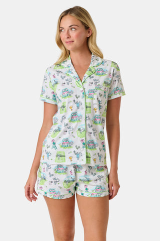 The Roller Rabbit Game, Set, Monkey Polo Pajamas is a two-piece set featuring a short-sleeve button-up shirt and matching shorts. Crafted from soft Pima Cotton, the white fabric is adorned with a playful cartoon animal and house print in green, blue, and yellow tones.