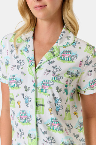 The Roller Rabbit Game, Set, Monkey Polo Pajamas is a two-piece set featuring a short-sleeve button-up shirt and matching shorts. Crafted from soft Pima Cotton, the white fabric is adorned with a playful cartoon animal and house print in green, blue, and yellow tones.