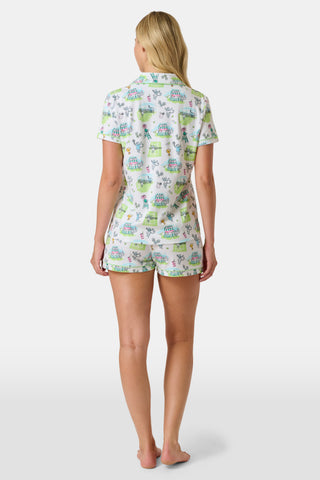 The Roller Rabbit Game, Set, Monkey Polo Pajamas is a two-piece set featuring a short-sleeve button-up shirt and matching shorts. Crafted from soft Pima Cotton, the white fabric is adorned with a playful cartoon animal and house print in green, blue, and yellow tones.