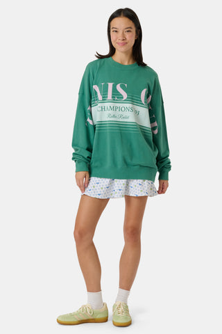 A person wearing a preppy green Roller Rabbit Tennis Champions Andie Sweatshirt stands in front of bookshelves. Only the lower part of their face is visible, and their hands are in their pockets.