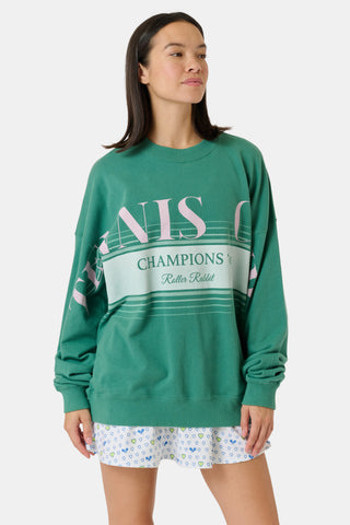 A person wearing a preppy green Roller Rabbit Tennis Champions Andie Sweatshirt stands in front of bookshelves. Only the lower part of their face is visible, and their hands are in their pockets.