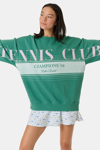 A person wearing a preppy green Roller Rabbit Tennis Champions Andie Sweatshirt stands in front of bookshelves. Only the lower part of their face is visible, and their hands are in their pockets.
