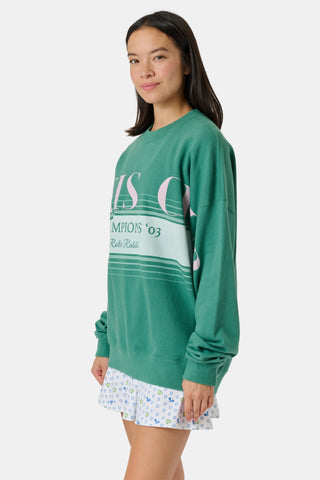 A person wearing a preppy green Roller Rabbit Tennis Champions Andie Sweatshirt stands in front of bookshelves. Only the lower part of their face is visible, and their hands are in their pockets.