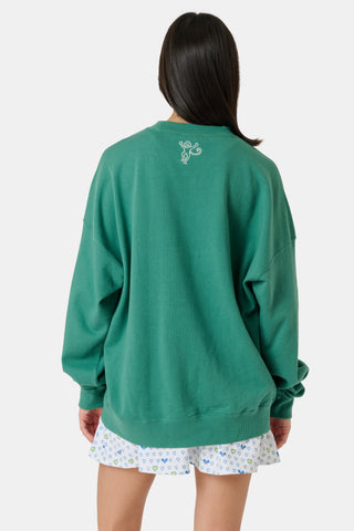 A person wearing a preppy green Roller Rabbit Tennis Champions Andie Sweatshirt stands in front of bookshelves. Only the lower part of their face is visible, and their hands are in their pockets.