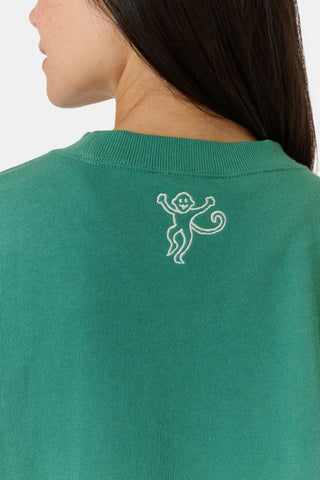 A person wearing a preppy green Roller Rabbit Tennis Champions Andie Sweatshirt stands in front of bookshelves. Only the lower part of their face is visible, and their hands are in their pockets.