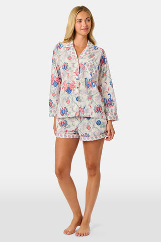 A woman stands against a plain white background, smiling slightly as she models the Amanda Leila Set from Roller Rabbit—a vintage-inspired cotton loungewear ensemble featuring a long-sleeve, button-up pajama top and matching shorts adorned with a colorful floral pattern.