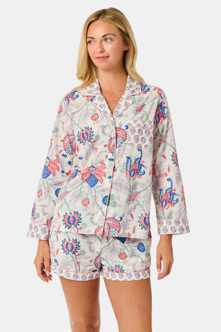 A woman stands against a plain white background, smiling slightly as she models the Amanda Leila Set from Roller Rabbit—a vintage-inspired cotton loungewear ensemble featuring a long-sleeve, button-up pajama top and matching shorts adorned with a colorful floral pattern.
