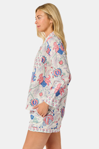A woman stands against a plain white background, smiling slightly as she models the Amanda Leila Set from Roller Rabbit—a vintage-inspired cotton loungewear ensemble featuring a long-sleeve, button-up pajama top and matching shorts adorned with a colorful floral pattern.