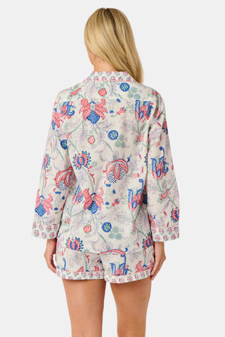 A woman stands against a plain white background, smiling slightly as she models the Amanda Leila Set from Roller Rabbit—a vintage-inspired cotton loungewear ensemble featuring a long-sleeve, button-up pajama top and matching shorts adorned with a colorful floral pattern.