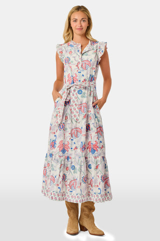 A person in the Amanda Nolan Dress by Roller Rabbit—a patterned, sleeveless, light-colored cotton maxi—stands on grass, holding out one side of the skirt. A wooden fence and lush greenery form the background; their face is not visible.