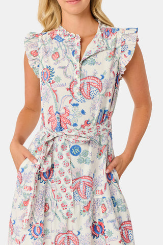 A person in the Amanda Nolan Dress by Roller Rabbit—a patterned, sleeveless, light-colored cotton maxi—stands on grass, holding out one side of the skirt. A wooden fence and lush greenery form the background; their face is not visible.