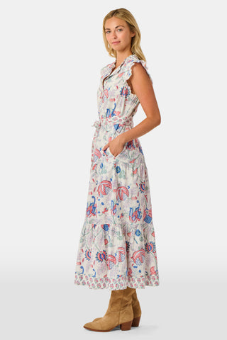 A person in the Amanda Nolan Dress by Roller Rabbit—a patterned, sleeveless, light-colored cotton maxi—stands on grass, holding out one side of the skirt. A wooden fence and lush greenery form the background; their face is not visible.