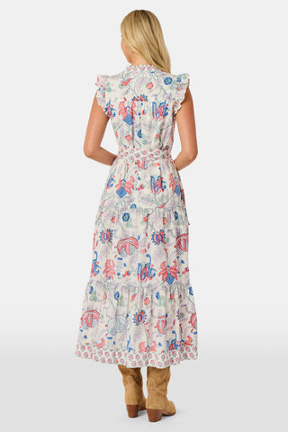 A person in the Amanda Nolan Dress by Roller Rabbit—a patterned, sleeveless, light-colored cotton maxi—stands on grass, holding out one side of the skirt. A wooden fence and lush greenery form the background; their face is not visible.