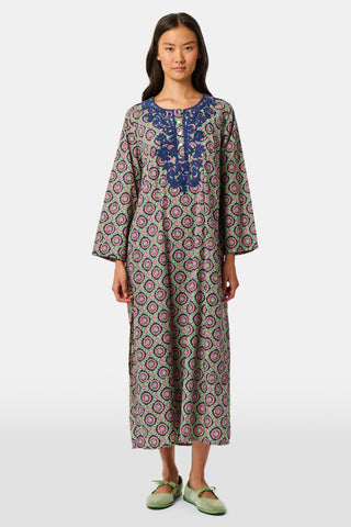 Wearing the Amelia Inez Long Kurta by Roller Rabbit, a woman with brown boots sits on the railing of a wooden porch, smiling, with a rustic building in the background.