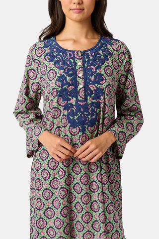 Wearing the Amelia Inez Long Kurta by Roller Rabbit, a woman with brown boots sits on the railing of a wooden porch, smiling, with a rustic building in the background.